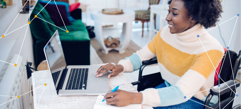 Young African American woman in a wheelchair working on her laptop and writing notes in a notebook at her home work desk